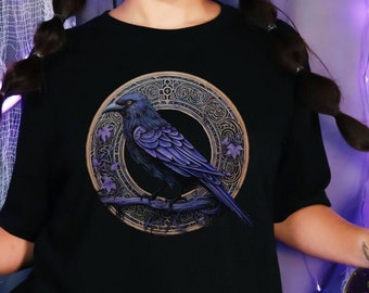 Puede incluir: Una camiseta negra con una ilustración de un cuervo morado dentro de un marco circular dorado con detalles intrincados. El cuervo está posado en una rama con hojas. La camiseta la lleva puesta una persona con el pelo largo y oscuro.