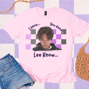 May include: A light pink t-shirt featuring a graphic of a person's face with the text "I know... You know... Lee Know..." in black and purple. The design includes a checkered pattern in the background.