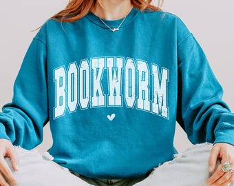 Puede incluir: Una sudadera azul turquesa con la palabra "BOOKWORM" impresa en blanco con un aspecto desgastado. La palabra está en una fuente grande y en negrita y está arqueada sobre un pequeño corazón blanco.