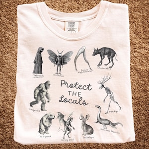 Puede incluir: Camiseta color crema con ilustraciones de críptidos como Mothman, Sasquatch y el Monstruo del Lago Ness. El texto "Protect the Locals" está impreso debajo de las ilustraciones. La camiseta es de material suave.