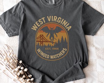 May include: Dark gray t-shirt featuring a circular graphic with the text "WEST VIRGINIA" and "WINGED WATCHERS". The graphic includes a bridge, a winged creature, trees, and the text "EST. 1966". The shirt is folded on a white patterned surface.