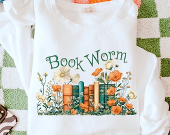 May include: White sweatshirt with the text "Book Worm" in green lettering. A stack of books with green spines and orange and white flowers are below the text.