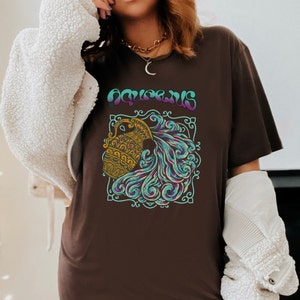 May include: Brown t-shirt with a graphic of the astrological sign Aquarius. The graphic features a stylized image of a water pitcher pouring water, surrounded by blue and purple swirls. The word "Aquarius" is written in purple and blue letters above the graphic.