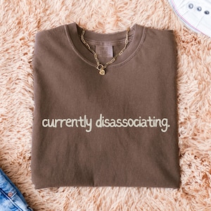 Puede incluir: Una camiseta marrón con la frase "currently dissociating" impresa en blanco. Un collar de cadena dorada descansa sobre la camiseta. La camiseta está doblada sobre una superficie suave de color melocotón.