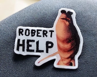 May include: A white patch with the words "ROBERT HELP" in black, featuring a cartoonish image of a creature. The creature is brown and black with a long, thin appendage. The patch is on a dark gray surface.