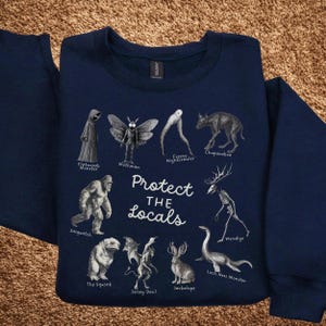 May include: Navy blue sweatshirt featuring illustrations of cryptids like Mothman, Sasquatch, and the Loch Ness Monster. The phrase "Protect the Locals" is printed in white. A unique and quirky design for fans of folklore and the unexplained.