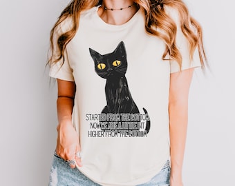 May include: A white t-shirt with a black cat illustration and the text "Started from the bottom now we are a little bit higher from the bottom".