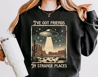 May include: Black sweatshirt with a vintage-style graphic of a UFO over a desert landscape. The text reads "I've got friends in strange places".