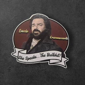 May include: A red and white vintage-style sticker featuring a portrait of a man with a beard and mustache. The text "Laszlo Cravensworth" and "She Speaks... The Bullshit!" is printed on the sticker.