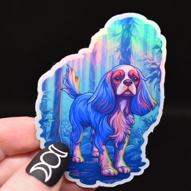 Może przedstawiać: Holograficzna naklejka przedstawiająca niebieskiego i r&oacute;żowego psa rasy Cavalier King Charles Spaniel stojącego w lesie. Pies ma długą, falującą sierść i patrzy bezpośrednio na widza.