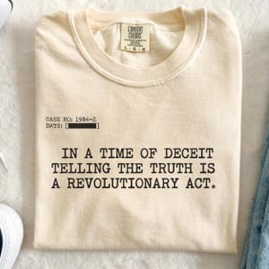 May include: Cream-coloured t-shirt with the text "IN A TIME OF DECEIT TELLING THE TRUTH IS A REVOLUTIONARY ACT." The shirt also has the text "CASE NO: 1984-Z DATE: [ ]" printed on it. The shirt has a crew neck and short sleeves.