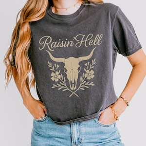 May include: Dark gray t-shirt with the text "Raisin' Hell" in a cursive font. The shirt features a beige bull skull graphic with floral and leaf accents. The shirt is made of a soft material.
