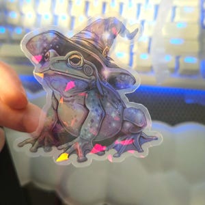 May include: A holographic sticker of a frog wearing a purple and blue wizard hat with stars and glitter. The frog is sitting on a pink and blue background.