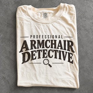 May include: A cream-colored short-sleeve t-shirt featuring the black text "PROFESSIONAL ARMCHAIR DETECTIVE" printed on the front. Below the text is a small black graphic of a magnifying glass. The shirt has a crew neckline and a brand label.