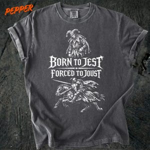 May include: Dark gray t-shirt with a distressed graphic featuring a jester and a knight on horseback. The text "BORN TO JEST FORCED TO JOUST" is displayed in a gothic font. The word "PEPPER" is in the upper left corner.
