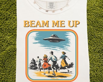 May include: White Comfort Colors t-shirt with a retro graphic featuring a UFO and people running away. The text "BEAM ME UP" and "This place sucks" are printed in a vintage style. A silver necklace with small charms is also visible.