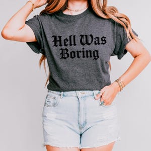 May include: A dark grey t-shirt featuring the text "Hell Was Boring" in a gothic font. The shirt is worn with light blue denim shorts. The outfit has a casual, contemporary style.