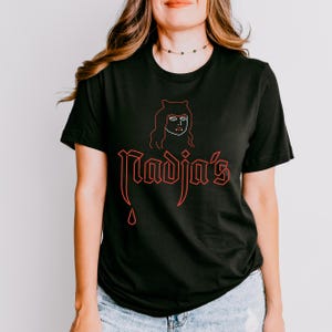 May include: Black t-shirt featuring a red graphic design of a woman with horns and the text "Nadja's" with a red blood drop dripping from the bottom.