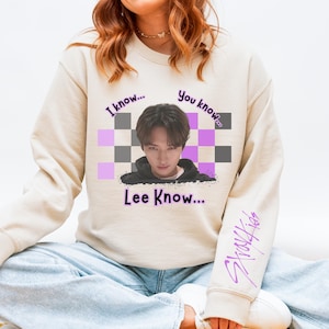 May include: Cream-coloured sweatshirt with a graphic of a person and the text "I know... You know... Lee Know...". The sleeve has the text "Stray Kids" in purple. The graphic includes a checkered pattern in grey and purple.