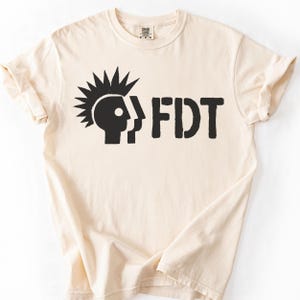 Puede incluir: Camiseta color crema con un gráfico negro. El gráfico presenta una cabeza estilizada con cresta y las letras "FDT". La camiseta tiene mangas cortas y cuello redondo.