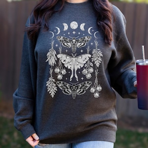 Puede incluir: Sudadera gris oscuro con un gráfico blanco de polillas, fases lunares y elementos florales. El diseño incluye tres polillas con intrincados patrones de alas, rodeadas de lunas crecientes y estrellas. La sudadera es de estilo casual y cómodo.