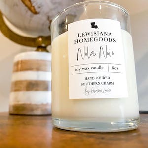May include: A clear glass candle jar with a white label that reads "LEWISIANA HOMEGOODS Nola Noir soy wax candle 6oz HAND POURED SOUTHERN CHARM by Andrew Lewis". The label also features a small image of the state of Louisiana.
