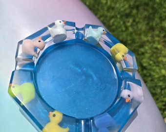 May include: A blue resin ashtray with a glittery center and a hexagonal shape. The ashtray is decorated with small, colorful duck figurines around the edges. The ducks are in various colors, including yellow, blue, and pink.