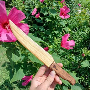 May include: A wooden knife with a light-colored blade and a dark brown handle. The handle has the initials "GW" carved into it. The knife is held in front of a background of pink flowers and green foliage.