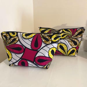 Peut inclure: Deux pochettes zippées arborant un motif géométrique vibrant en blanc, rose vif, jaune et noir. Le tissu présente des formes stylisées rappelant des pétales et des lignes courbes disposées en motif répétitif, avec une fermeture éclair noire.