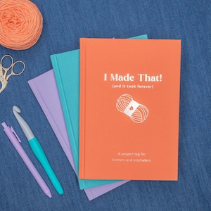 May include: An orange project log book with the text "I Made That!" and a yarn ball illustration. The book is stacked with other colorful notebooks, a crochet hook, and pens. A ball of orange yarn and scissors are also visible.