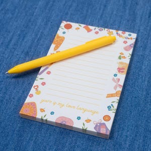 May include: A notepad with a decorative border featuring yarn-related illustrations and the text "yarn is my love language." A yellow pen rests on the lined pages. The notepad is on a blue denim surface.