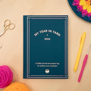 May include: A teal bullet journal titled "MY YEAR IN YARN 2026" with white text and a decorative border. The journal lies on a wooden surface, accompanied by a yellow pen, a pink crochet hook, scissors, and yarn balls.
