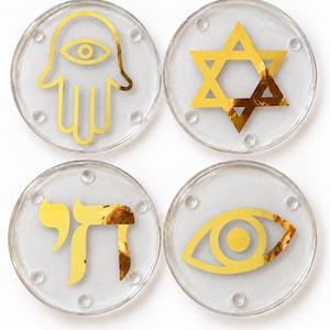 May include: A set of four circular clear coasters, each featuring a distinct gold foil design. The symbols include a hamsa hand, a Star of David, the Hebrew word Chai, and an eye. Each coaster has small raised circular feet on the surface.