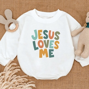 May include: A white long-sleeved baby romper featuring the text 'JESUS LOVES ME' printed in a multi-colored, retro-style font with shades of teal, orange, and olive green. The garment has a round neckline and elasticized leg openings.