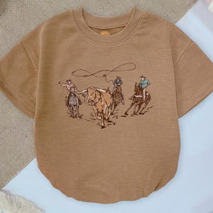 May include: A tan short-sleeved t-shirt featuring a graphic print of three cowboys on horseback roping a longhorn steer. The shirt has a crew neckline and a relaxed fit, showcasing a detailed illustration of western ranch life in brown tones.