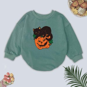 May include: A teal-colored baby bodysuit with a crew neck and long sleeves. The front features a graphic of a black cat resting on a carved orange pumpkin with green leaves. The design is in a vintage Halloween style.