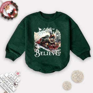 May include: A dark green sweatshirt with long sleeves. The front features a detailed illustration of a vintage train with the word "BELIEVE" in white script. The train is set against a snowy winter scene, with Santa's sleigh in the sky.