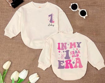 May include: Two cream-colored long-sleeved shirts with pink and purple designs. One shirt says "In My 1 Year Old Era" and the other says "1 Aubrey".