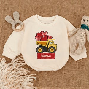 May include: Cream-colored baby romper with a yellow dump truck graphic carrying red hearts. The name "William" is printed on a red rectangular label below the truck. A wooden bunny ear teether and a teddy bear are next to the romper.
