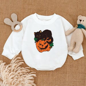 May include: White baby onesie with a Halloween-themed graphic. The graphic features a black cat resting on top of an orange jack-o'-lantern. The onesie has long sleeves and a round neckline. A wooden bunny-shaped teether and a stuffed bear are also in the image.