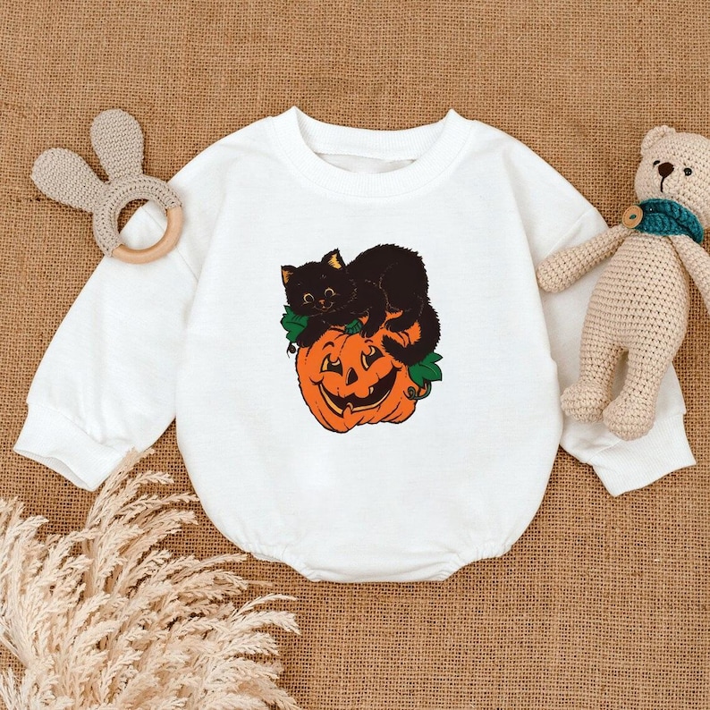 May include: White baby onesie with a Halloween-themed graphic. The graphic features a black cat resting on top of an orange jack-o'-lantern. The onesie has long sleeves and a round neckline. A wooden bunny-shaped teether and a stuffed bear are also in the image.