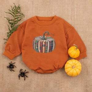 May include: An orange baby romper with a pumpkin design made of books. Two small yellow pumpkins and two black plastic spiders are next to the romper. A sprig of green leaves is in the upper left corner.