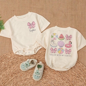 May include: Two white short-sleeved shirts with a pink bow and the name "Sophia" on one shirt and a pink flamingo, watermelon, and other summer-themed designs on the other shirt. The text on the second shirt reads "Chillin with my floaties".