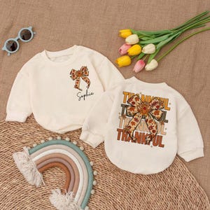 May include: Two cream-colored sweatshirts with long sleeves. One features a bow graphic and the name "Sophia". The other has a bow graphic and the word "THANKFUL" in stacked text. The sweatshirts are on a woven mat with a rainbow and flowers.