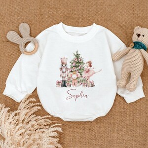 May include: White baby romper with a Christmas-themed design. The design includes a nutcracker, Christmas tree, ballerina, and the name "Sophia" in a script font. A wooden bunny teether and a stuffed teddy bear are also in the image.
