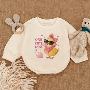 May include: Cream-colored baby romper with long sleeves. The front features a graphic of a yellow chick wearing sunglasses, a pink bow, and a bandana, blowing a bubble. The text "ONE CUTE CHICK" is printed above the chick. A bunny-shaped teether and a teddy bear are on either side.