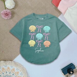 May include: A teal green short-sleeved t-shirt with a graphic of colorful seashells and bows. The name "Sophia" is printed below the graphic.