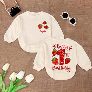 May include: Two cream-colored long-sleeved shirts with strawberry graphics. One shirt says "Sophia" and the other says "Berry 1st Birthday".
