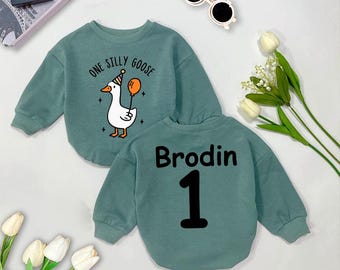 May include: A teal blue long-sleeve sweatshirt with the text "One Silly Goose" and a cartoon goose with a party hat and a balloon on the front. The back of the sweatshirt has the name "Brodin" and the number "1" in black.