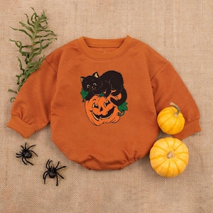 May include: An orange baby romper with a black cat and jack-o'-lantern graphic. The long-sleeved outfit has a crew neck and is decorated with a smiling pumpkin and a black cat. Two small pumpkins and plastic spiders are also in the image.